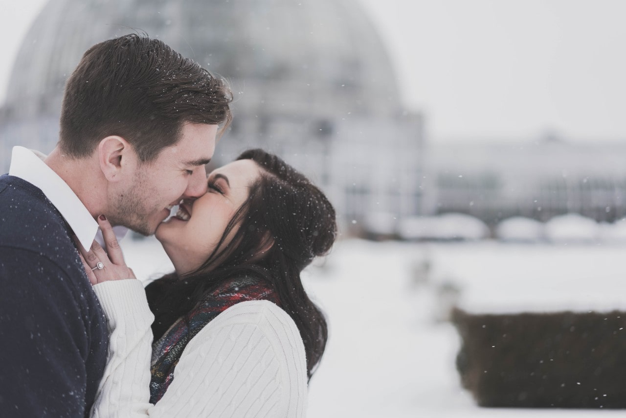 A couple embraces with smiling faces close, set against a snowy backdrop and blurred dome structure.