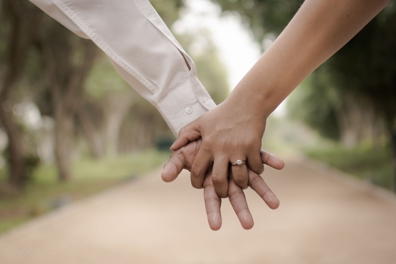 Two people hold hands on a blurred pathway, one wearing a white shirt and the other a yellow gold solitaire engagement ring.