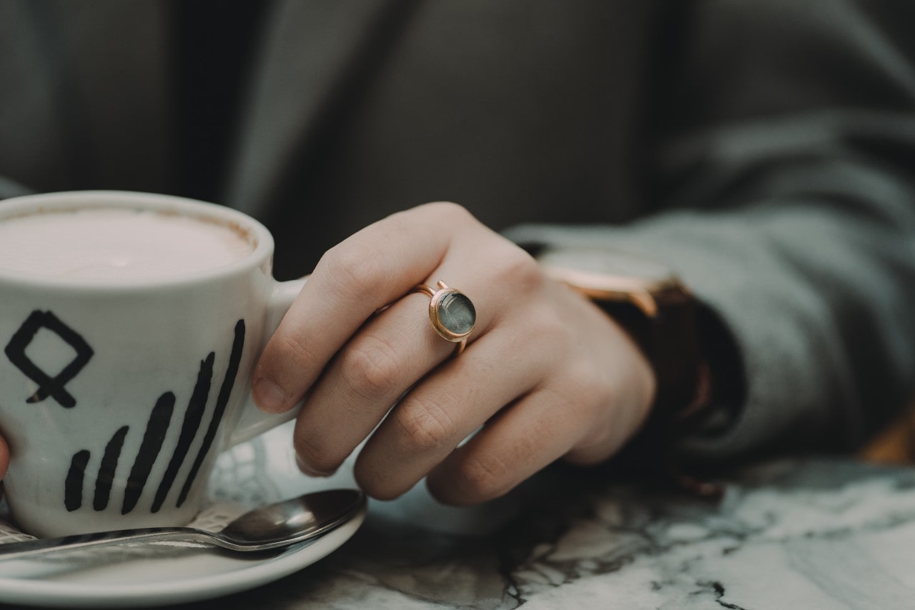 A close up of a hand with a gold ring with a round, translucent stone holding a ceramic mug of coffee