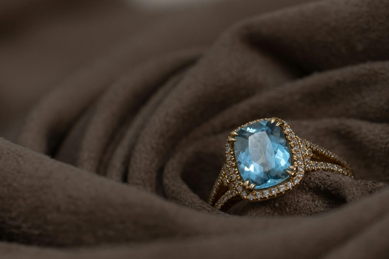 A gold ring with a large, sparkling blue gemstone surrounded by small diamonds, placed on soft brown fabric.